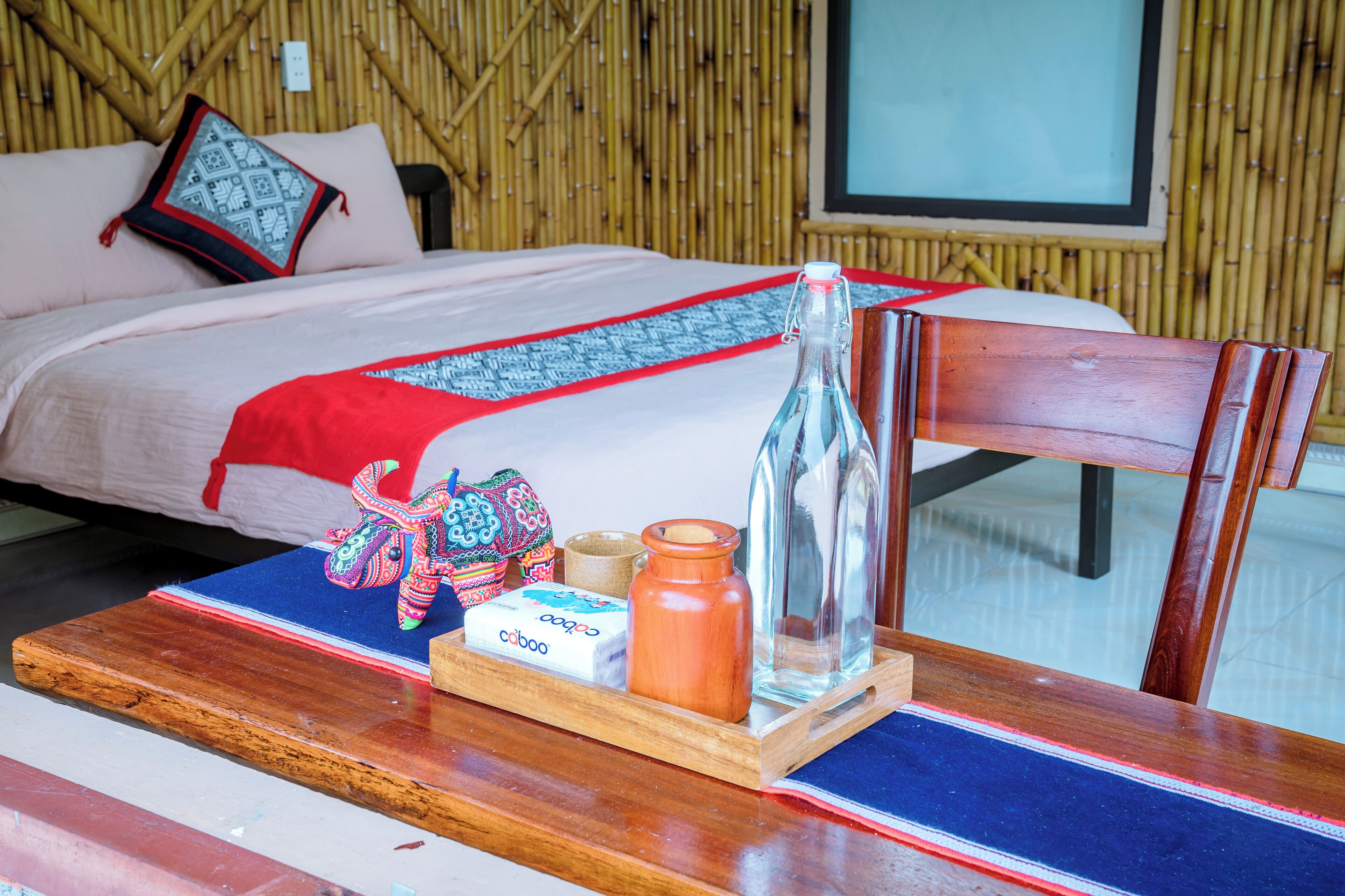 Un lit recouvert d’une couvre-lit aux motifs rouges et bleus, une table de chevet en bois avec une bouteille, une bougie et un sac en tissu tressé.
