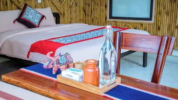 Un lit recouvert d’une couvre-lit aux motifs rouges et bleus, une table de chevet en bois avec une bouteille, une bougie et un sac en tissu tressé.