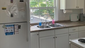 Une cuisine avec des meubles blancs, un réfrigérateur, une fenêtre donnant sur un espace verdoyant, un évier et un plan de travail.