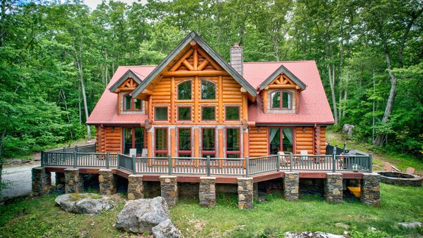A log cabin with a stone foundation, a wooden deck, and a red roof. - Big Elk Lodge - Luxury home in Eagles Nest, walk to Great Camp, firepit (Banner Elk)