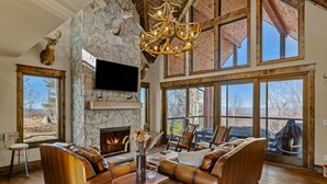Amplia sala de estar con chimenea de piedra, sofás de cuero y ventanas grandes que ofrecen vistas al exterior.