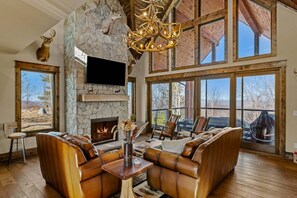 A spacious living room with a stone fireplace, leather sofas, and large windows offering a view of the outdoors. - Safe Haven at Eagles Nest - views, hot tub, firepit, fireplace, game table (Banner Elk)