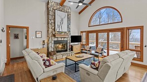 A spacious living room with a stone fireplace, two sofas, a television, and large windows offering a view of the outdoors. - Daisy Ridge - Expansive Mountain View, Pets OK, Beech Mtn Club option, foosball, arcade (Beech Mountain)
