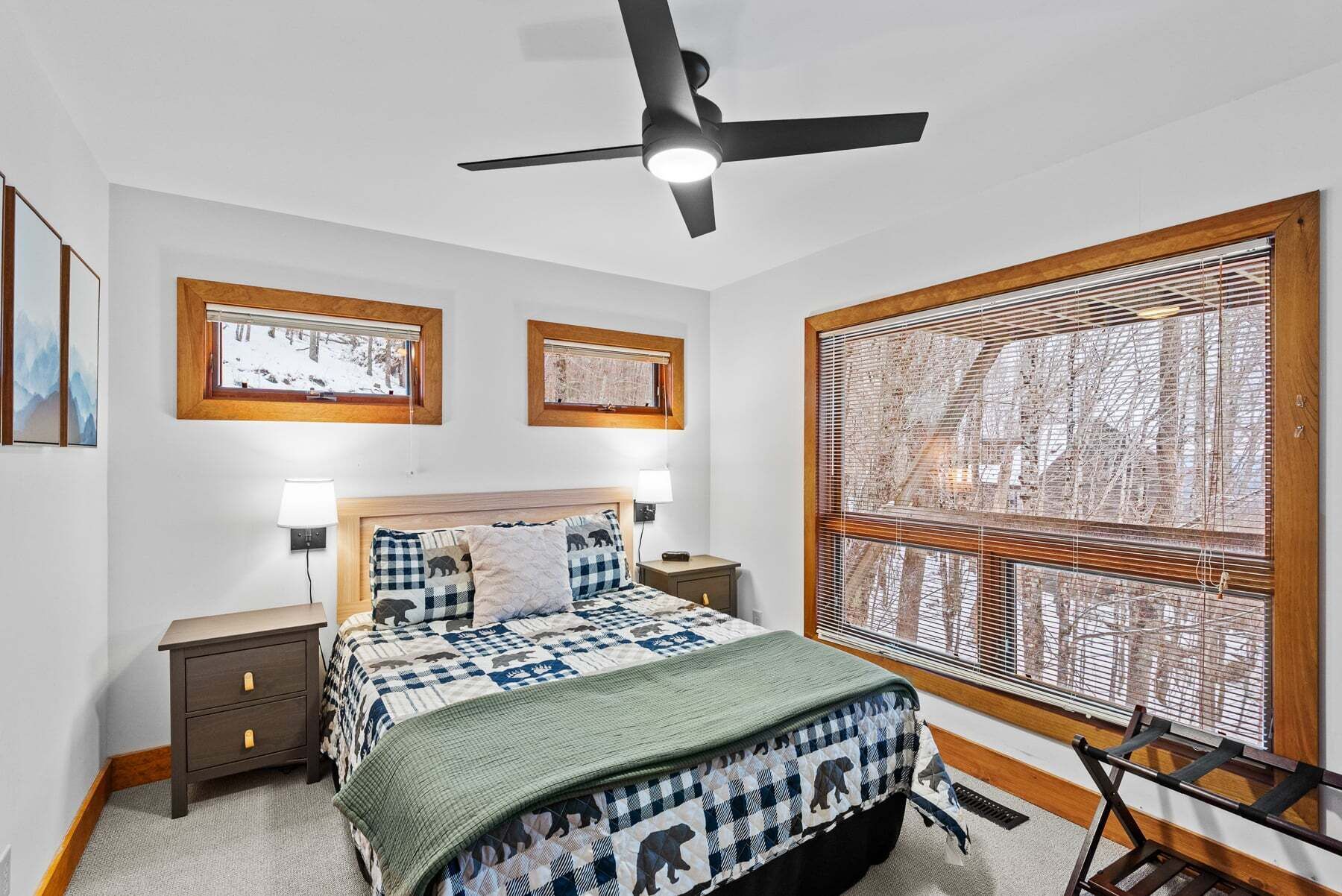 Ein Schlafzimmer mit Bett, Nachttischen, Deckenventilator, zwei Fenstern mit Jalousien und Bildern an den Wänden.