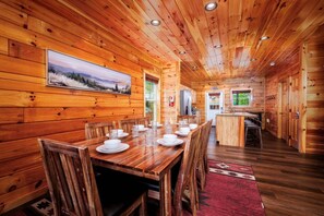 A wooden cabin interior with a dining area, kitchen, and a large window. - Fiddler's Friend - Hot Tub Soaks, Theater Movie Nights & Game Room Competitions (Pigeon Forge)