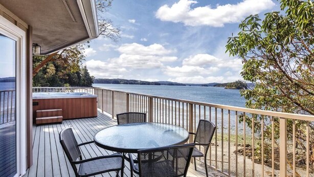 Una terraza con mesa y sillas de vidrio, un jacuzzi y vistas a un lago.