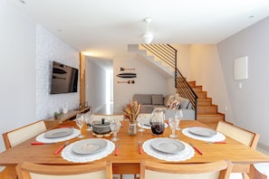 Un coin repas avec une table en bois dressée pour quatre personnes, un vase contenant des fleurs séchées et un escalier menant à un autre niveau.