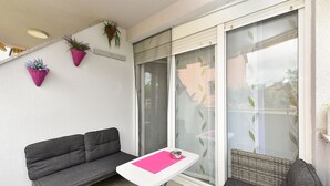 Un balcon avec un canapé gris, une petite table surmontée d’un tapis rose et deux chaises en osier.