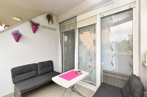 Un balcon avec un canapé gris, une petite table surmontée d’un tapis rose et deux chaises en osier.