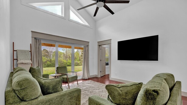 A spacious living room with a large window, a ceiling fan, a flat-screen TV, and a green sofa.