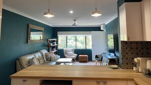 A modern living room with a wooden coffee table, a sofa, a chair, and a dining table. There is a window with a view of trees outside. - OP 63 - Rest & Recharge at Your Beachside Escape (Coffs Harbour)
