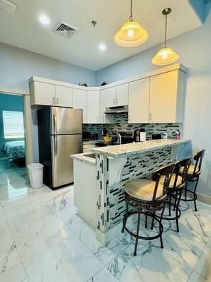 A modern kitchen with white cabinets, a stainless steel refrigerator, a marble countertop, and a bar area with chairs. - Marisol Sunset Villa #154 (South Padre Island)