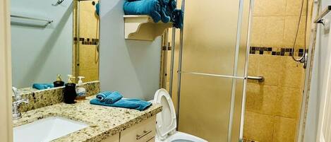 Ein Badezimmer mit Waschbecken, Spiegel, Toilette und Dusche. Auf der Ablage liegt ein blaues Handtuch.