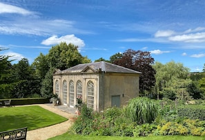 A small, stone building with arched windows, surrounded by a well-maintained garden with various plants and a lawn. - South Cottage - Sherborne, Dorset (Sherborne)