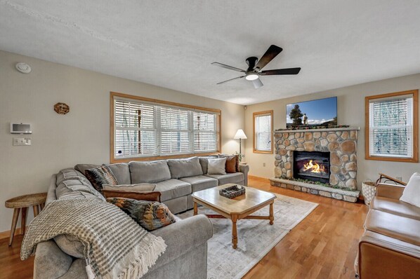 A living room with a stone fireplace, a ceiling fan, and large windows with blinds. - Ski/family/kids/fully-stocked kitchen/spacious (Coolbaugh Township)