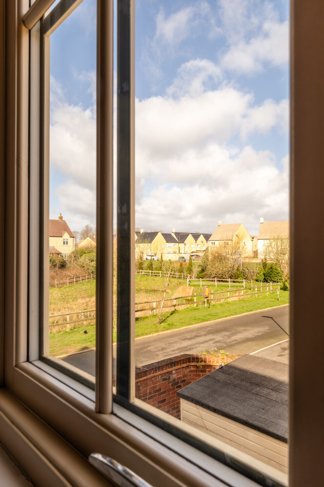 Une vue à travers une fenêtre donnant sur un quartier résidentiel, avec des maisons, un champ d’herbe et un mur de briques.