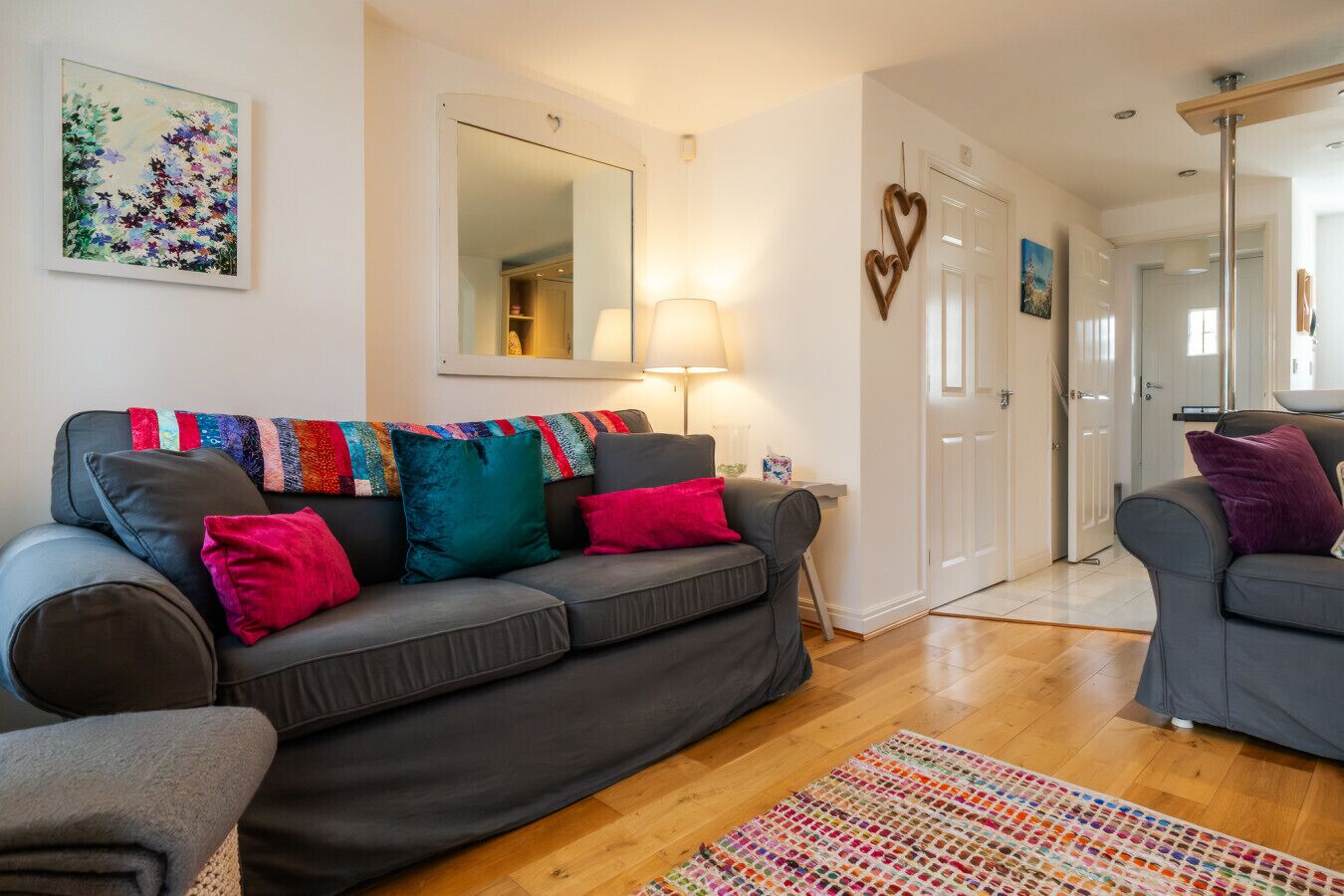 A living room with a grey sofa, colorful throw pillows, a mirror, a lamp, and a heart-shaped decoration on the wall.