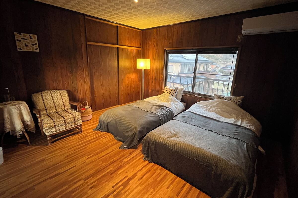 Une pièce avec un plancher en bois, deux lits avec une literie grise, une chaise en bois, une lampe et une fenêtre donnant sur un paysage.