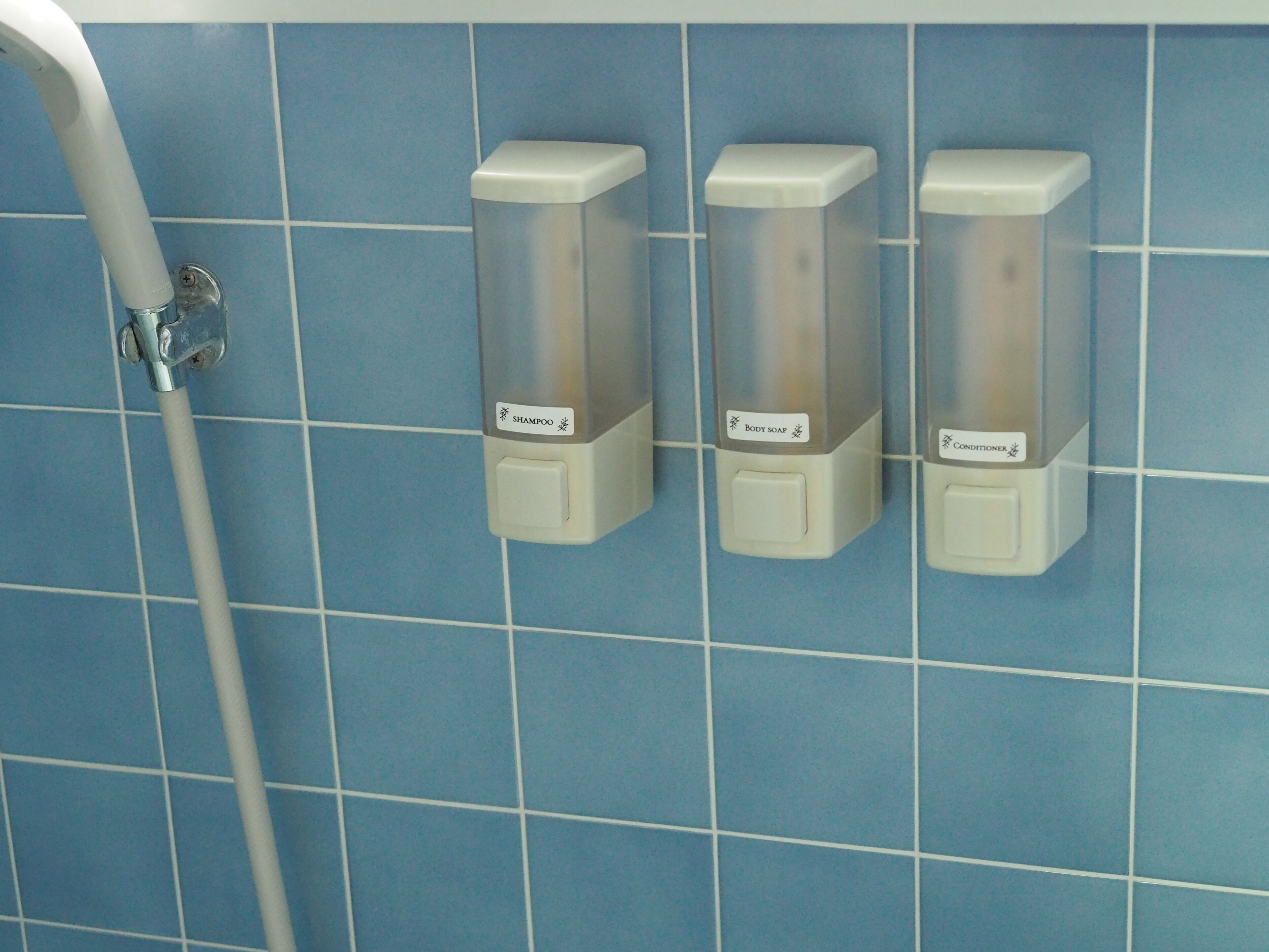 A bathroom with blue tiled walls, three dispensers labeled for shampoo, body soap, and conditioner, and a showerhead.