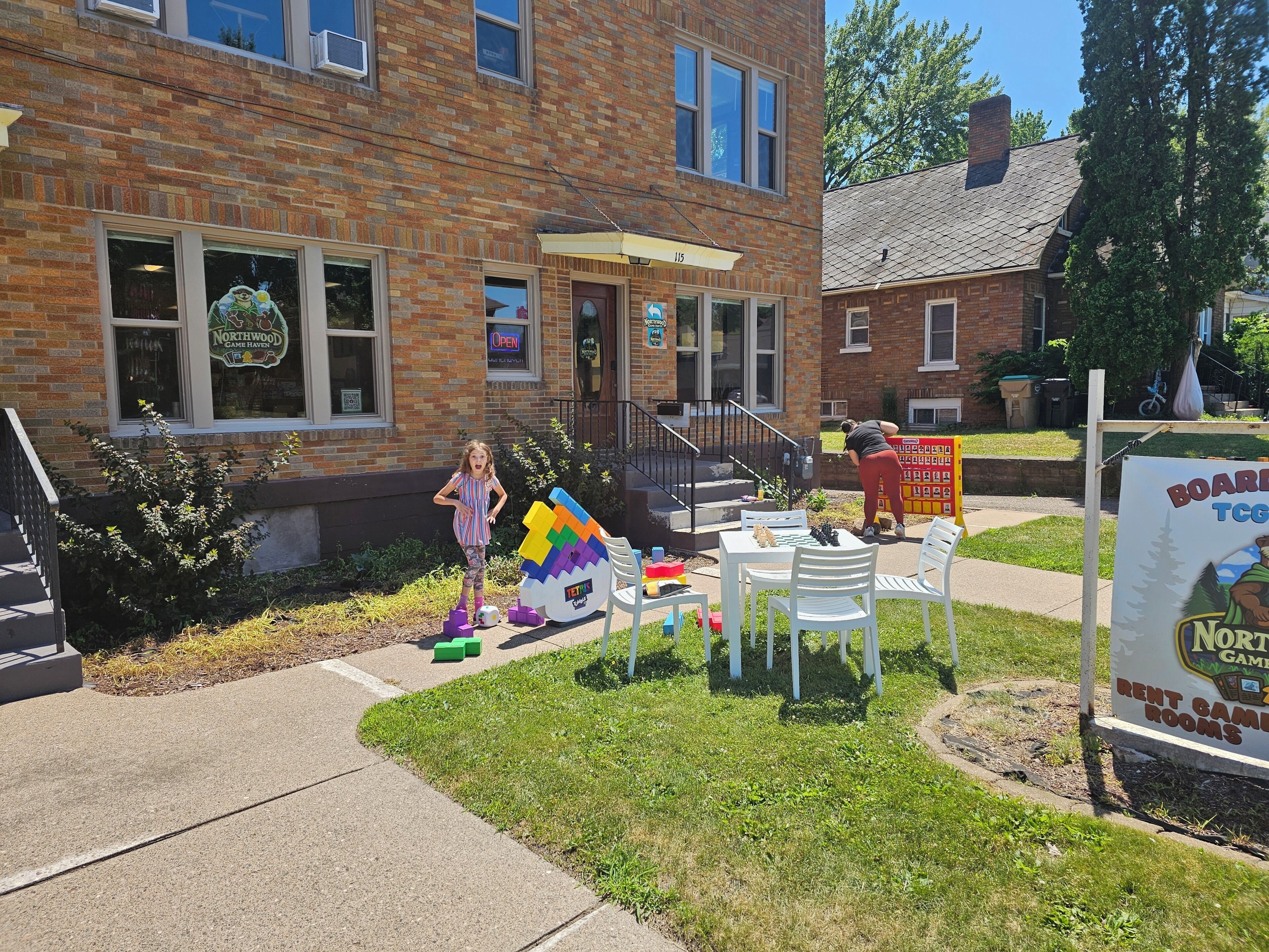 A brick building with a sign for a game room. There are outdoor tables and chairs, and a child playing with toys.