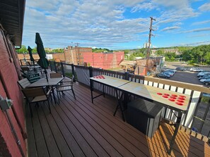Una terraza de madera con mobiliario exterior, mesa de ping-pong y vista a un estacionamiento y edificios.