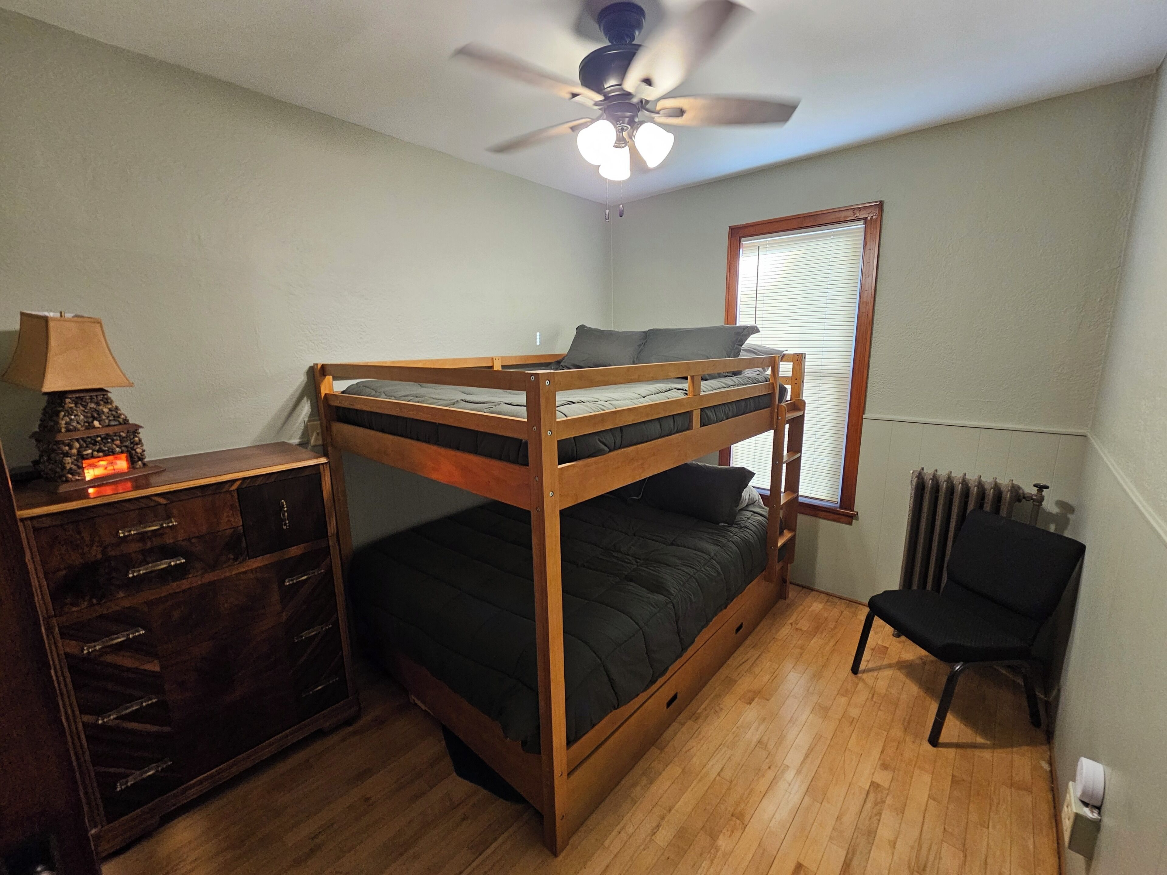 Une chambre avec un lit superposé, une commode en bois, une lampe de bureau, un radiateur et un ventilateur de plafond.