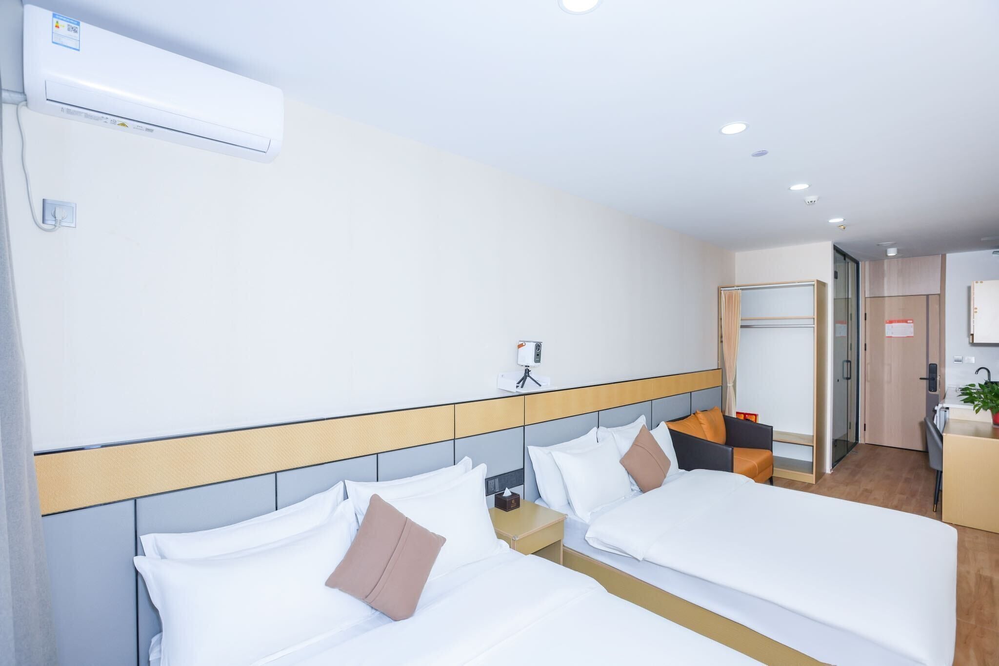 A hotel room with two beds, a bedside table, a nightstand, a mirror, a wall-mounted air conditioner, a small desk with a chair, and a kitchenette.