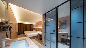 Una habitación de hotel moderna con una cama grande, un baño independiente con lavamanos y espejo, y un ventanal amplio.