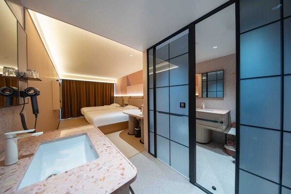 A modern hotel room with a large bed, a separate bathroom with a sink and mirror, and a large window.