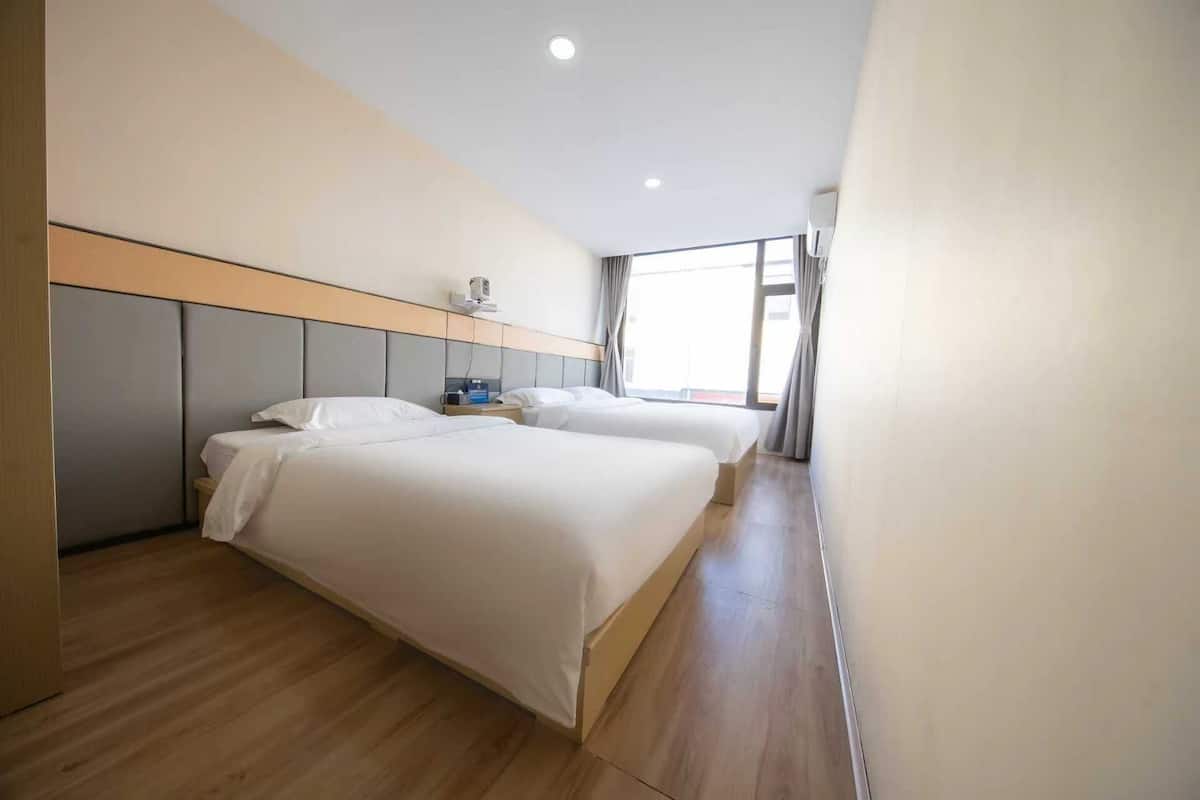 Hotellihuone, jossa on kaksi sänkyä, puulattia, sängynpääty, ikkuna verhoilla ja kattovalaisin.