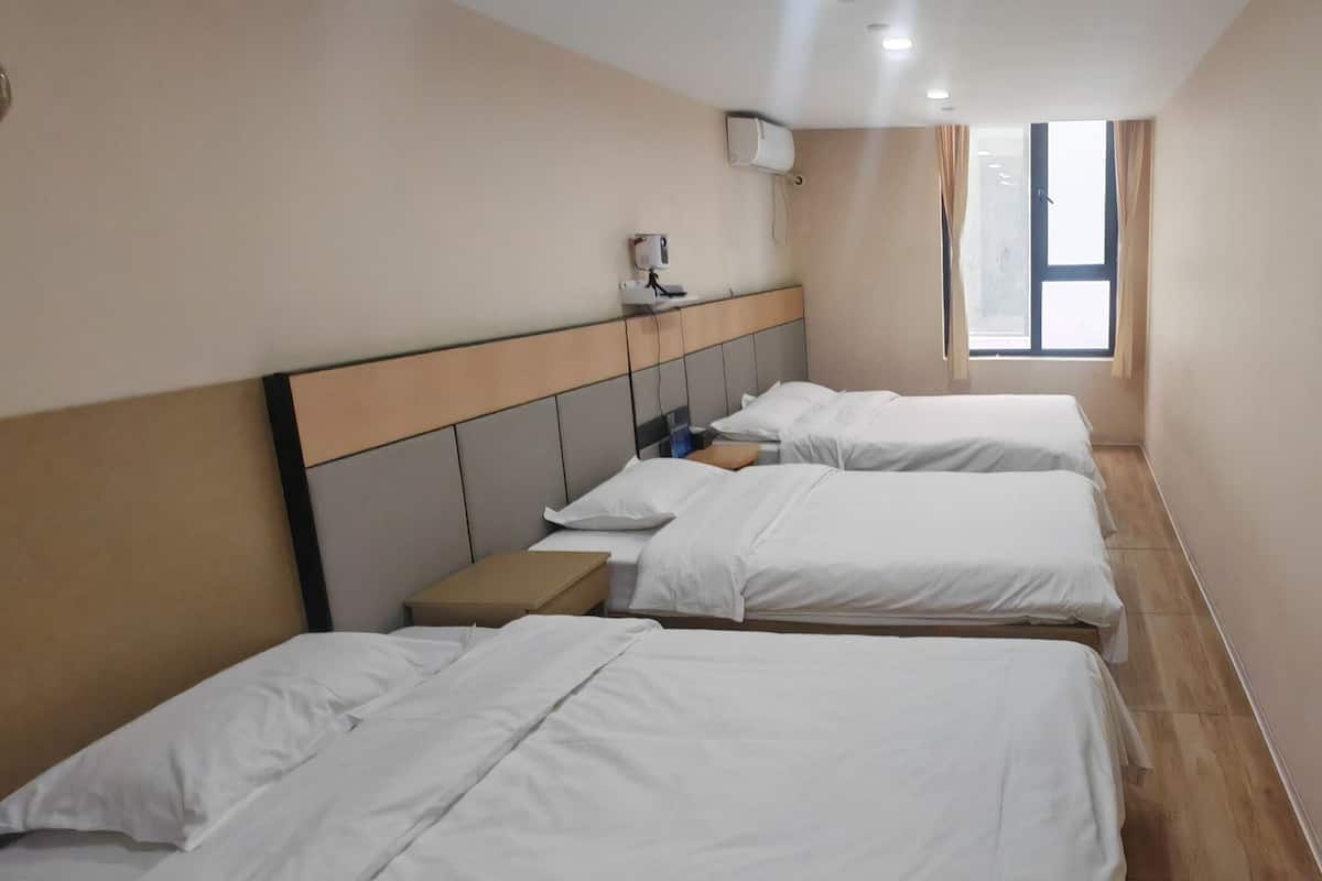 Hotellihuone, jossa on kolme sänkyä, yöpöytä, ikkuna ja seinälle asennettu ilmastointilaite.