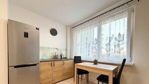 A compact kitchen with a refrigerator, wooden cabinets, a dining table with chairs, a clock, and a window with sheer curtains. - Bystrzycka Comfort Apartment (Wrocław)