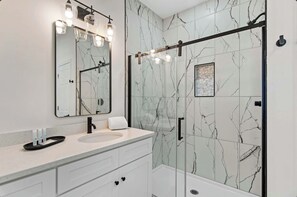Ein modernes Badezimmer mit Marmorfliesenspiegel, einem groĂen, beleuchteten Spiegel, einer Glasdusche und einem weiĂen Waschtisch mit Schubladen.
