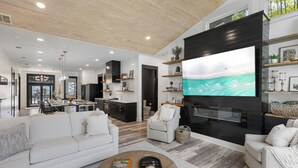 A modern living room with a large flat-screen TV, a white sofa, a coffee table, and a kitchen in the background. - Poole Party! Incredible Pool, Beachside of 30A (Seagrove Beach)