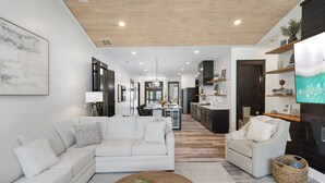A modern living room with a sofa, armchair, coffee table, and a television. There is a kitchen in the background with cabinets and a dining area. - Poole Party! Incredible Pool, Beachside of 30A (Seagrove Beach)