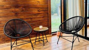 Une pièce aux murs en bois, deux chaises au design épuré en fil de fer noir, une petite table ronde avec une bougie, et une porte vitrée donnant sur l’extérieur.
