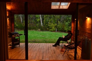 Une terrasse en bois avec un barbecue, une personne assise sur une chaise, et une vue sur une cour verdoyante avec des arbres.