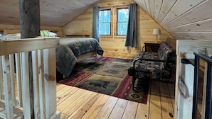 A cozy attic bedroom with wooden floors, a bed, a chair, and a window with curtains. - Cozy Woodland Cabin Living with Loft and Brookside Views (Boscawen)