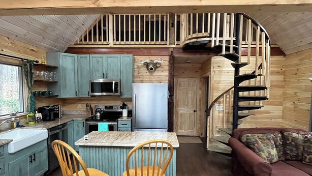 A cabin interior with a kitchen, dining area, and a spiral staircase. - Cozy Woodland Cabin Living with Loft and Brookside Views (Boscawen)