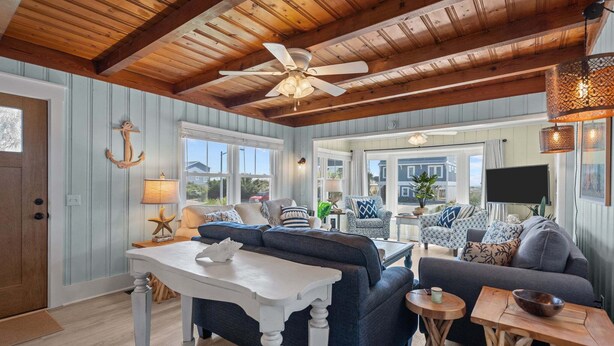 A spacious living room with a ceiling fan, wooden beams, a large window, and a cozy seating area.