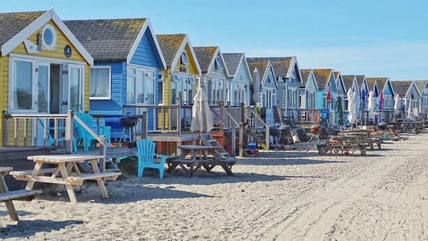 A row of beach huts with wooden benches and tables, a sandy beach, and a clear sky. - Modern town house situated in a quiet location with views across the harbour (Christchurch)