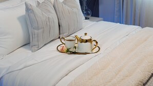 Un lit parfaitement fait, avec du linge de lit blanc, un service à thé orné de motifs dorés et une rose décorative posée sur un plateau.