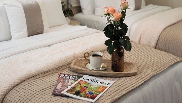 A neatly made bed with white bedding, a vase with flowers, a tray with a cup and saucer, and a magazine on the bed.