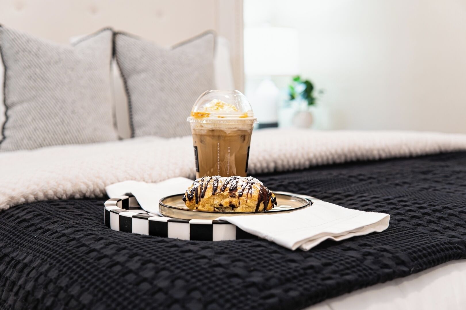 A bed with a black and white striped pillow, a black knitted blanket, a white pillow, and a glass of iced coffee with a pastry on a plate.