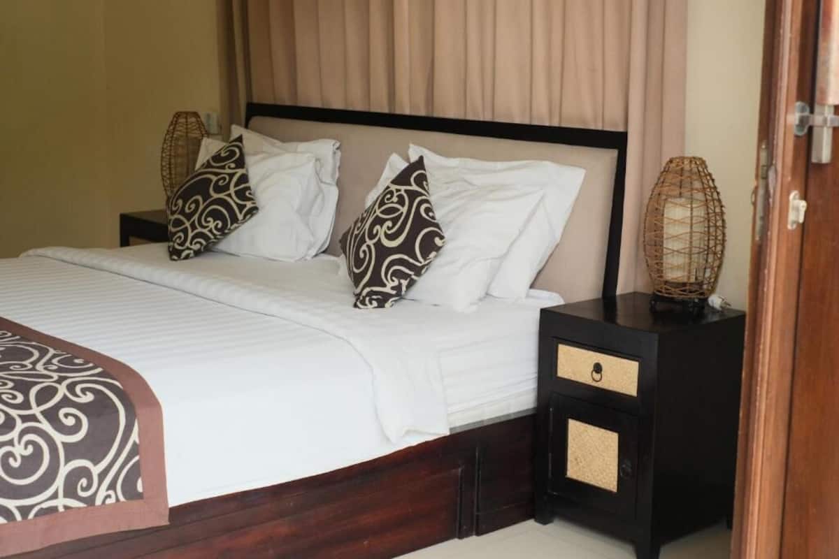 A neatly made bed with a patterned bedspread, two pillows with a swirl design, a black nightstand with a drawer, and a woven lamp.