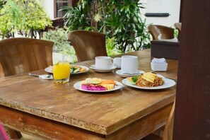 A wooden dining table with plates of food, a glass of orange juice, and white cups. - Umah Gatra Ubud (Ubud)