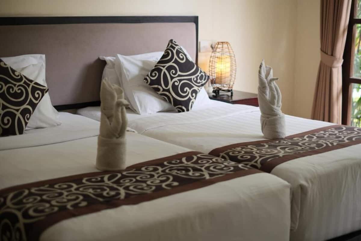 A hotel room with two beds, decorative pillows, a bedside lamp, and a towel rack with towels.