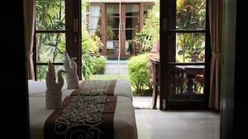 A hotel room with a bed, a view of the garden through open doors, and a dining area visible through the glass doors.