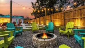 Een achtertuin met een vuurkorf, omringd door groene Adirondack-stoelen, een houten omheining en lichtjes.