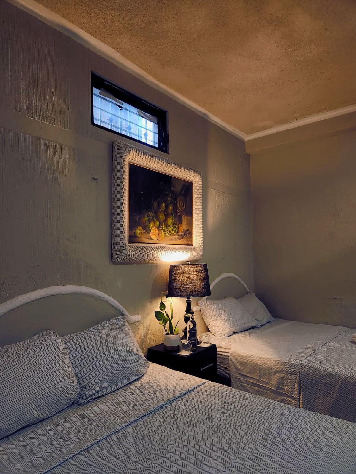 A hotel room with two beds, a nightstand with a lamp, a framed painting, and a window.